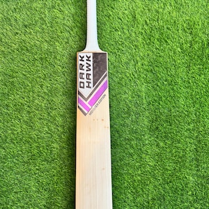 May include: A wooden cricket bat with a white grip and the text "DARK HAWK" in black and white. The bat has purple and silver accents and the words "MONSTER EDITION". The bat is on a green grass background.