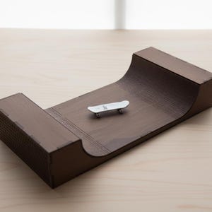 Op de afbeelding: Een miniatuur houten skateboard ramp in donkerbruin, met een klein wit skateboard dat op het gebogen oppervlak rust. De helling heeft een getextureerde zijkant en een soepele, gebogen overgang. De achtergrond is een licht houten oppervlak.