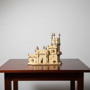 Pu&ograve; includere: Un modello in legno tagliato al laser di un castello dettagliato, con pi&ugrave; torri, finestre ad arco e intricati dettagli architettonici. Il modello &egrave; di colore beige chiaro e poggia su un tavolo di legno marrone scuro.