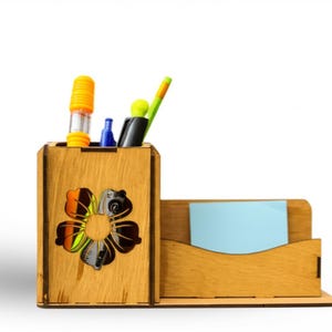 May include: A wooden desk organizer with a flower cutout design. The organizer has a section for pens and markers, and a separate section for sticky notes. The organizer is made of light brown wood and contains pens and a marker.