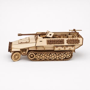 May include: A laser-cut wooden model of a half-track armored vehicle. The model features a front wheel, a rear track system, a mounted cannon, and a small machine gun at the rear. The light-colored wood construction shows intricate details.