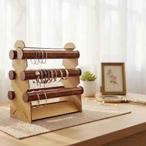 May include: A three-tiered wooden jewelry display stand with brown dowels and light-colored supports. The stand holds various bracelets. The stand is on a beige woven mat on a wooden table. A mirror and framed picture are in the background.