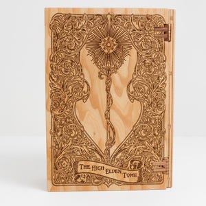 Puede incluir: Un libro de madera con intrincados diseños cortados con láser. La portada presenta un emblema central con una estrella y un báculo, rodeado de adornos ornamentados. El texto "THE HIGH ELDEN TOME" está grabado en una pancarta. Bisagras de color cobre aseguran el libro.