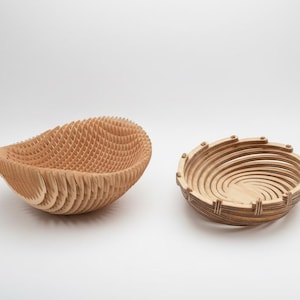 Puede incluir: Dos cuencos de madera. Uno es un cuenco en forma de lágrima, de color marrón claro, con un interior en forma de panal. El otro es un cuenco redondo con un diseño en espiral y una serie de anillos de madera. Ambos cuencos están sobre un fondo blanco.
