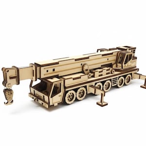 May include: A wooden model of a mobile crane, with a detailed design including a boom, hook, and multiple wheels. The crane is made from light-coloured wood, showing the natural grain and texture. The model is a construction-themed item.