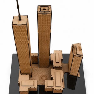 Può includere: Un modellino architettonico in legno tagliato al laser del complesso del World Trade Center, caratterizzato da due torri alte con facciate a griglia, una piazza centrale con linee radiali incise e diversi edifici circostanti più piccoli.
