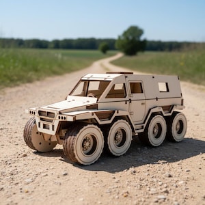 Puede incluir: Maqueta de madera de un vehículo de seis ruedas, construida con madera de color claro. El modelo presenta neumáticos, ventanas y un techo detallados. El vehículo está situado en un camino de tierra, con un fondo borroso de árboles y cielo azul.