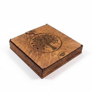 Puede incluir: Caja de madera rectangular con tapa con bisagras, con un diseño de árbol tallado. La caja tiene un acabado marrón oscuro y el árbol está dentro de un marco circular con detalles de hojas.