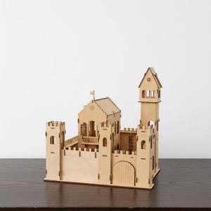 May include: A wooden castle model with multiple towers, a gate, and a flag. The model is made of light-colored wood and sits on a dark surface. The castle has detailed architectural features.