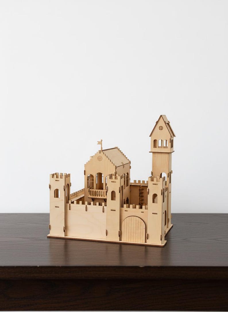 May include: A wooden castle model with multiple towers, a gate, and a flag. The model is made of light-colored wood and sits on a dark surface. The castle has detailed architectural features.