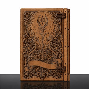 Peut inclure: Une couverture de livre rectangulaire en bois avec des détails gravés au laser. Le motif présente un bâton central surmonté d'une étoile, entouré de feuillages et de fleurs. Une bannière en ruban vide est située en bas.