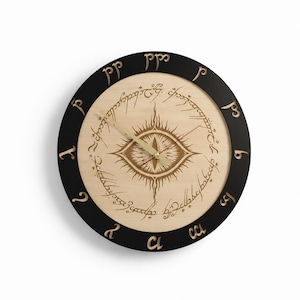 Op de afbeelding: Een ronde wandklok met een zwarte buitenrand voorzien van goudkleurige Tengwar-tekens. De lichtgekleurde houten wijzerplaat toont een centraal oogmotief omringd door een cirkelvormige inscriptie in een schrijflettertype, met goudkleurige wijzers.
