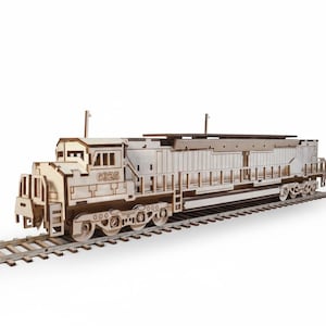 Puede incluir: Maqueta de madera de una locomotora diésel sobre una vía. El modelo es de madera clara y presenta cortes detallados, incluido el número "G325" en el lateral. El tren está sobre un fondo blanco.