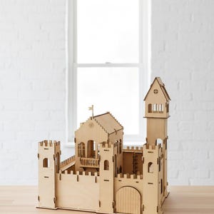 May include: A wooden castle model with a detailed design. The castle features multiple towers, walls, and a gate. The model is made of light-colored wood and sits on a wooden surface. A window is visible in the background.