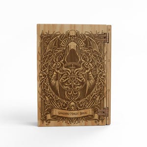 May include: Wooden book with intricate laser-cut design. The cover features a hooded figure, skull, and ornate details. The banner reads "Shadow Magic Book". Hinges are visible on the right side. The book is made of light-colored wood.
