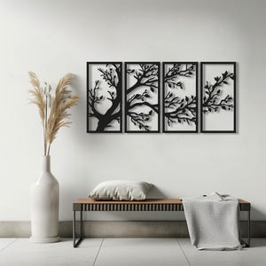 Puede incluir: Arte mural de metal negro de cuatro paneles que representa ramas y hojas de árboles estilizadas. La obra de arte está enmarcada en negro y montada en una pared blanca. Un jarrón blanco con plantas secas, un banco, una almohada y una manta están en primer plano.