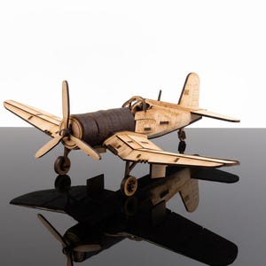 Könnte beinhalten: Ein Holzmodellflugzeug mit Propeller und Flügeln. Das Modell besteht aus hellem Holz mit einem dunkelbraunen Rumpf. Das Flugzeug steht auf einer reflektierenden Oberfläche.