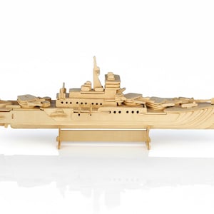 May include: A detailed wooden model of a warship, constructed from light-coloured wood. The model showcases the ship's design, including turrets, a bridge, and other features. It is presented against a white backdrop, highlighting the craftsmanship.