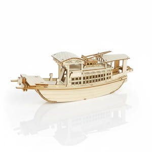 Puede incluir: Maqueta de madera de un barco tradicional con detalles intrincados. El barco presenta un casco curvo, múltiples cubiertas y tallas decorativas. El modelo está hecho de madera de color claro y se muestra sobre una superficie blanca.