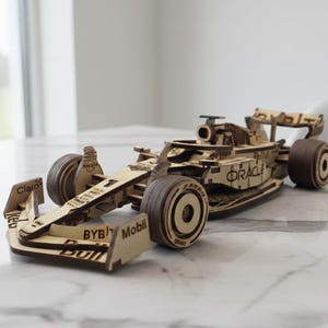 Puede incluir: Maqueta de madera de un coche de carreras de Fórmula 1. El coche es de madera marrón claro con ruedas marrones oscuras. El modelo tiene logotipos de patrocinadores y detalles.