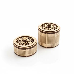 Puede incluir: Dos cajas decorativas de madera con tapas. Las cajas son cilíndricas con un acabado marrón claro y detalles en marrón oscuro. Las tapas presentan un diseño floral recortado. Las cajas están sobre un fondo blanco.
