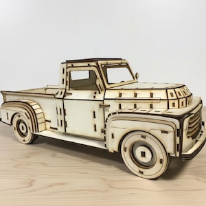 Op de afbeelding: Een houten model van een vintage pick-up truck. De truck is gemaakt van lichtgekleurd hout met donkerdere houten accenten, die de details van het voertuig laten zien, inclusief de cabine, laadbak en wielen. Het model staat op een lichtgekleurd houten oppervlak.
