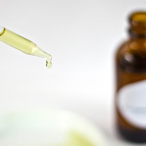 May include: A glass dropper with a yellow liquid dripping from the tip. The dropper is in focus, while a brown glass bottle with a white label is out of focus in the background.