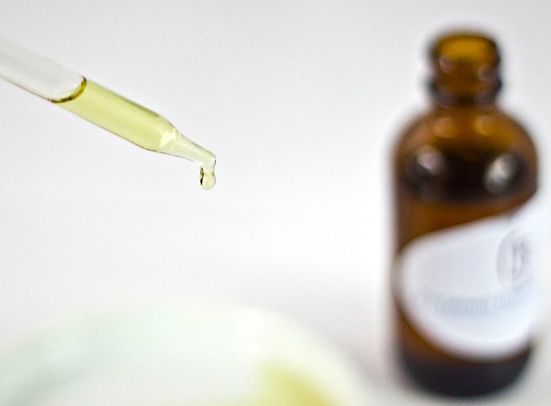 May include: A glass dropper with a yellow liquid dripping from the tip. The dropper is in focus, while a brown glass bottle with a white label is out of focus in the background.