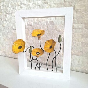 Puede incluir: Una obra de arte mural enmarcada en blanco con flores de amapola amarillas con centros y tallos negros. Las flores son tridimensionales y parecen estar hechas de papel o un material similar. El marco es rectangular y está pintado de blanco.