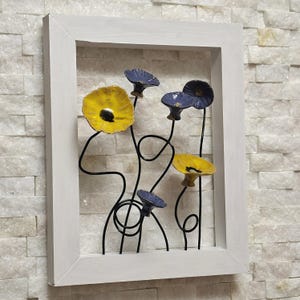 Op de afbeelding: Een ingelijst kunstwerk voor aan de muur met gestileerde metalen bloemen. De bloemen zijn geel en blauw met zwarte stelen, tegen een witte bakstenen achtergrond. De lijst is wit.