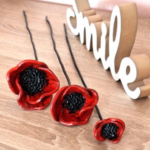 Può includere: Tre steli di fiori di papavero rossi con centri e steli neri. I fiori sono disposti su una superficie di legno, con un cartello decorativo "smile" sullo sfondo.