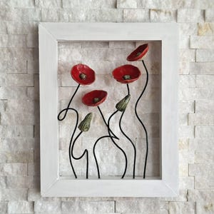 Könnte beinhalten: Gerahmtes Wandkunstwerk mit roten Mohnblumen mit schwarzen Stielen und grünen Knospen. Die Mohnblumen sind vor einem weißen Ziegelhintergrund platziert und in einem weißen Holzrahmen eingeschlossen. Das Kunstwerk hat einen 3D-Effekt.
