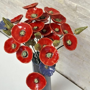 May include: A ceramic bouquet of red poppy flowers with green and blue accents. The poppies have a glossy finish and are arranged in a grey vase. The flower petals are a vibrant red with green centres.