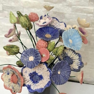 Könnte beinhalten: Ein Strauß aus handgefertigten Keramikblumen an dünnen grünen Drahtstielen. Die Blüten zeigen verschiedene Texturen und Farben in Blau, Weiß, Rosa und Grün, ergänzt durch gesprenkelte Muster und kleine, akzentuierte Blütenmitten an jedem einzelnen Exemplar.