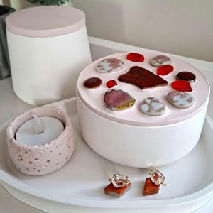 Op de afbeelding: Een ronde witte container met een lichtroze deksel versierd met diverse kleurrijke keramische en glazen fragmenten. Daarnaast staat een kleine getextureerde lichtroze kaarsenhouder met een wit waxinelichtje en twee kristallen oorbellen.
