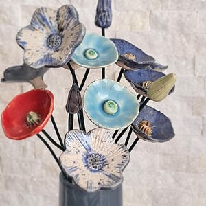 Könnte beinhalten: Eine Ansammlung von Keramikblumen an dünnen schwarzen Metallstielen in verschiedenen Farben wie Rot, Hellblau, Dunkelblau und Weiß mit blauen Sprenkeln. Jede Blume hat eine ausgeprägte Mitte, einige zeigen geschlossene Keramikknospen.