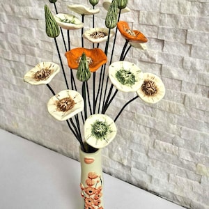 Puede incluir: Un arreglo floral de cerámica con flores de amapola naranjas, blancas y verdes. Las flores están en un jarrón crema con un diseño floral. Los tallos son negros.
