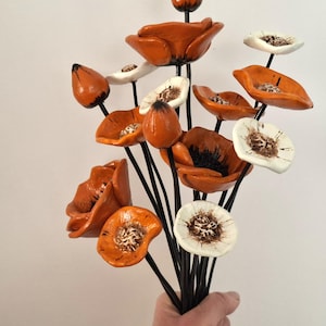 Könnte beinhalten: Ein Strauß aus orangefarbenen und weißen Keramik-Mohnblumen. Die Blumen haben detaillierte Zentren und sind auf schwarzen Stielen angeordnet. Der Strauß wird vor einem schlichten Hintergrund gehalten.
