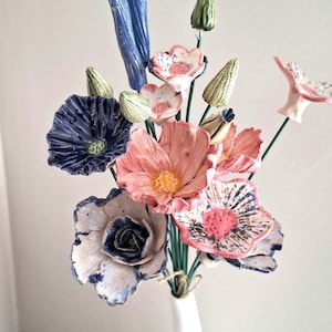 Può includere: Un bouquet di fiori in ceramica realizzati a mano con un mix di petali blu scuro, rosa tenue e bianchi con dettagli maculati. La composizione include diversi boccioli in ceramica verde su steli sottili, raccolti in un vaso bianco.