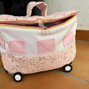 Könnte beinhalten: Eine Stoff-Reisetasche in Form eines Wohnwagens. Die Tasche ist weiß mit rosa Blumen- und Tupfenakzenten. Sie hat einen gelben Reißverschluss, einen Griff und kleine schwarz-weiße Räder. Die Tasche ist geöffnet und zeigt den Inhalt.