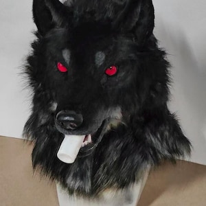 May include: A black wolf mask with red eyes and a white object in its mouth. The mask has a realistic fur texture and is mounted on a white stand. The wolf's expression is fierce.