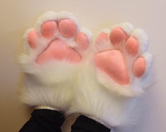 May include: A pair of white furry paw gloves with pink paw pads. The gloves are designed to resemble animal paws, with the pink pads contrasting against the white fur. The gloves are worn by a person wearing black clothing.