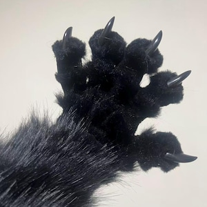May include: A black furry paw with five sharp, black claws. The paw is made of soft, plush material and has long, black fur extending from the wrist area. The claws are pointed and glossy.