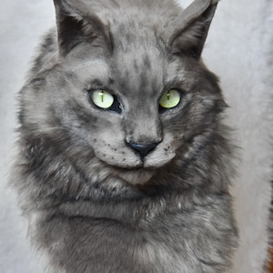 Pode incluir: Uma escultura realista de cabeça de gato com pelo cinzento denso e texturizado, orelhas pontiagudas e olhos verdes brilhantes e marcantes. O rosto felino tem um nariz escuro, bigodes visíveis e uma expressão detalhada e realista.