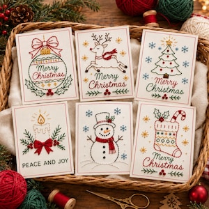 May include: Six white greeting cards featuring embroidered holiday designs including a Christmas ornament, a reindeer, a tree, a candle with the text PEACE AND JOY, a snowman, and a stocking, all with the text Merry Christmas.