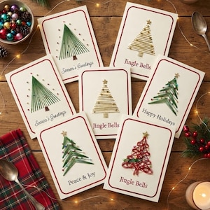 Può includere: Un set di sei biglietti d'auguri rettangolari con disegni di alberi di Natale ricamati in filo verde, oro e rosso. Ogni biglietto è bordato di rosso e riporta scritte come Season's Greetings, Jingle Bells, Happy Holidays e Peace & Joy.
