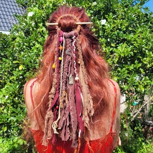 Puede incluir: Cabello rojo peinado con una horquilla de madera y adornado con tela y detalles de cuentas. El cabello está peinado en un moño con mechones de tela marrón, burdeos y crema, acentuados con cuentas y abalorios. La mujer lleva una camiseta roja.