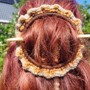 Puede incluir: Un envoltorio de moño de pelo de ganchillo en tonos marrones, beige y dorados, asegurado con una horquilla de madera. El envoltorio circular se coloca alrededor del pelo castaño rojizo de una persona, con la horquilla de madera sujetando el pelo en su sitio. El fondo es una escena soleada al aire libre.