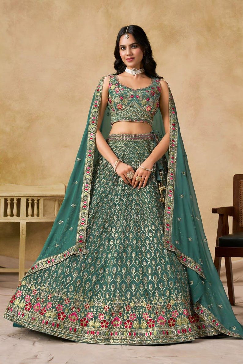 May include: A teal green formal dress with intricate gold embroidery and floral accents. The dress features a crop top, a long skirt, and a matching sheer shawl. The design includes a detailed pattern and is suitable for special occasions.