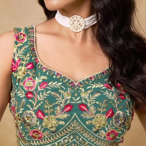 May include: Emerald green sleeveless top with intricate floral embroidery in pink, gold, and green. The top features a scoop neckline and is adorned with sequins. A white beaded choker necklace with a gold pendant completes the look. This is a traditional Indian garment.